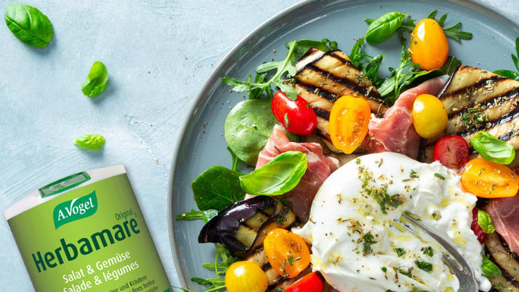 A. Vogel Herbamare seasoning next to a vibrant salad with grilled aubergine, cherry tomatoes, prosciutto, fresh greens, and burrata cheese.