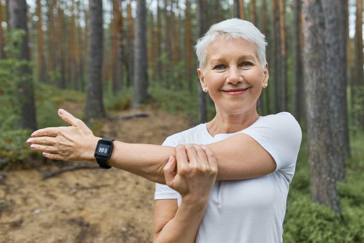 Smiling short-haired woman in a white t-shirt stretches her arm outdoors in a forest, promoting bone health with supplements and activity tracking.