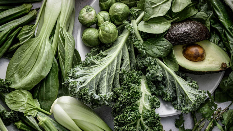 Assorted fresh green vegetables including kale, spinach, brussels sprouts, and avocado, representing vegan supplements by The Good Stuff.